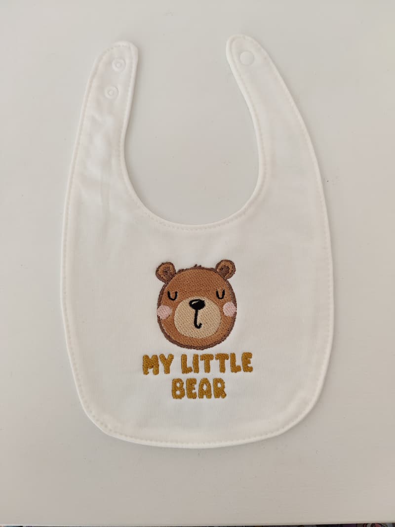 Fotografia di un bavaglino per neonati bianco con ricamato sopra la testa di un orsetto marrone e la scritta "My Little Bear" in marrone.