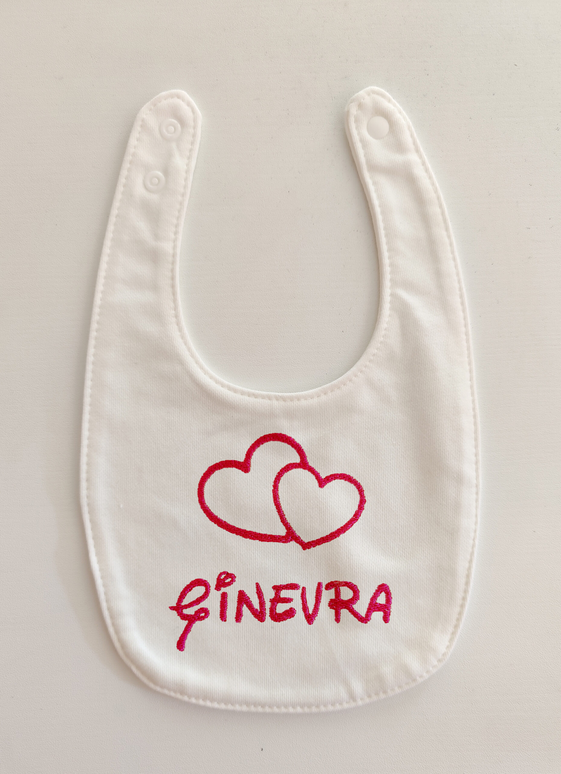 Fotografia di un bavaglino per neonati bianco con ricamato in rosso il nome "Ginevra" e due cuori uno accanto all'altro.