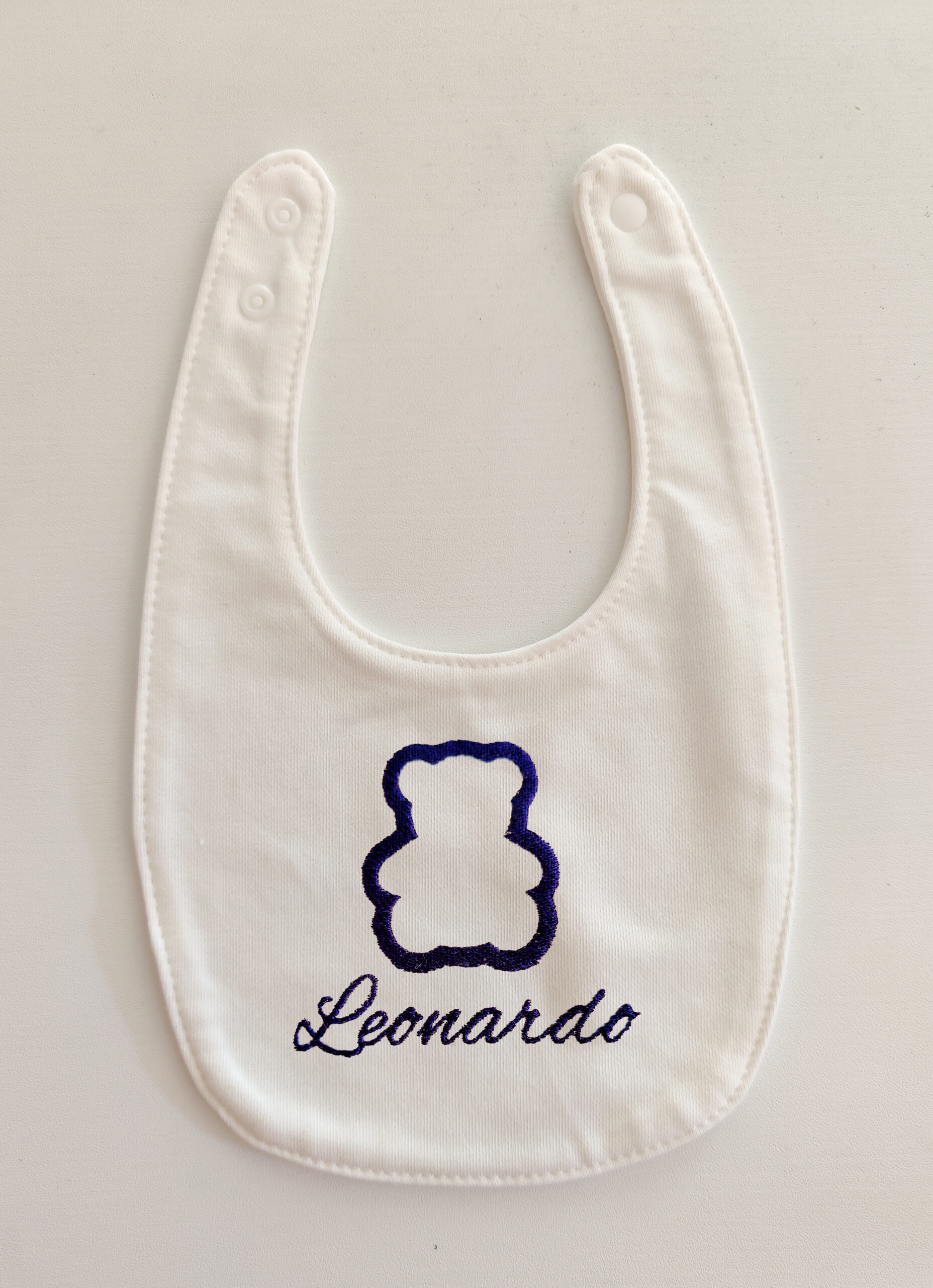 Fotografia di un bavaglino bianco per neonato con ricamato il nome "Leonardo" in blu con la sagoma di un orsetto.
