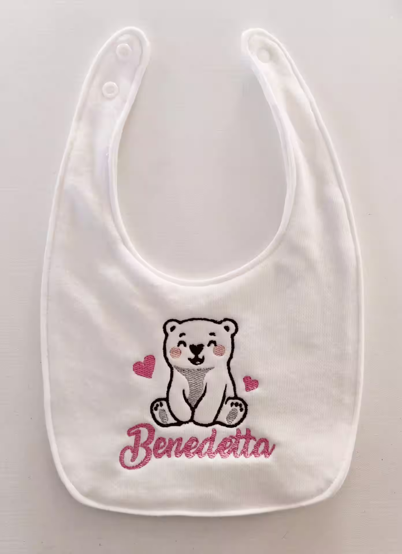 Fotografia di un bavaglino bianco con ricamato un orso polare con dei cuori ed il nome "Benedetta" in rosa.