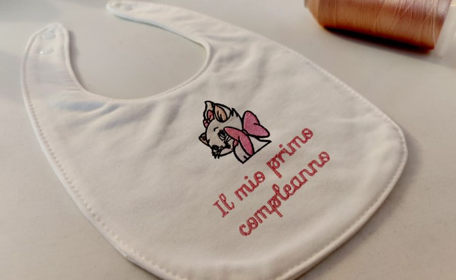 Fotografia di un bavaglino per neonati messo in obliquo con sopra ricamato la faccia di Minou, una delle protagoniste del cartone animato "Gli Aristogatti" e la scritta "Il mio primo compleanno".